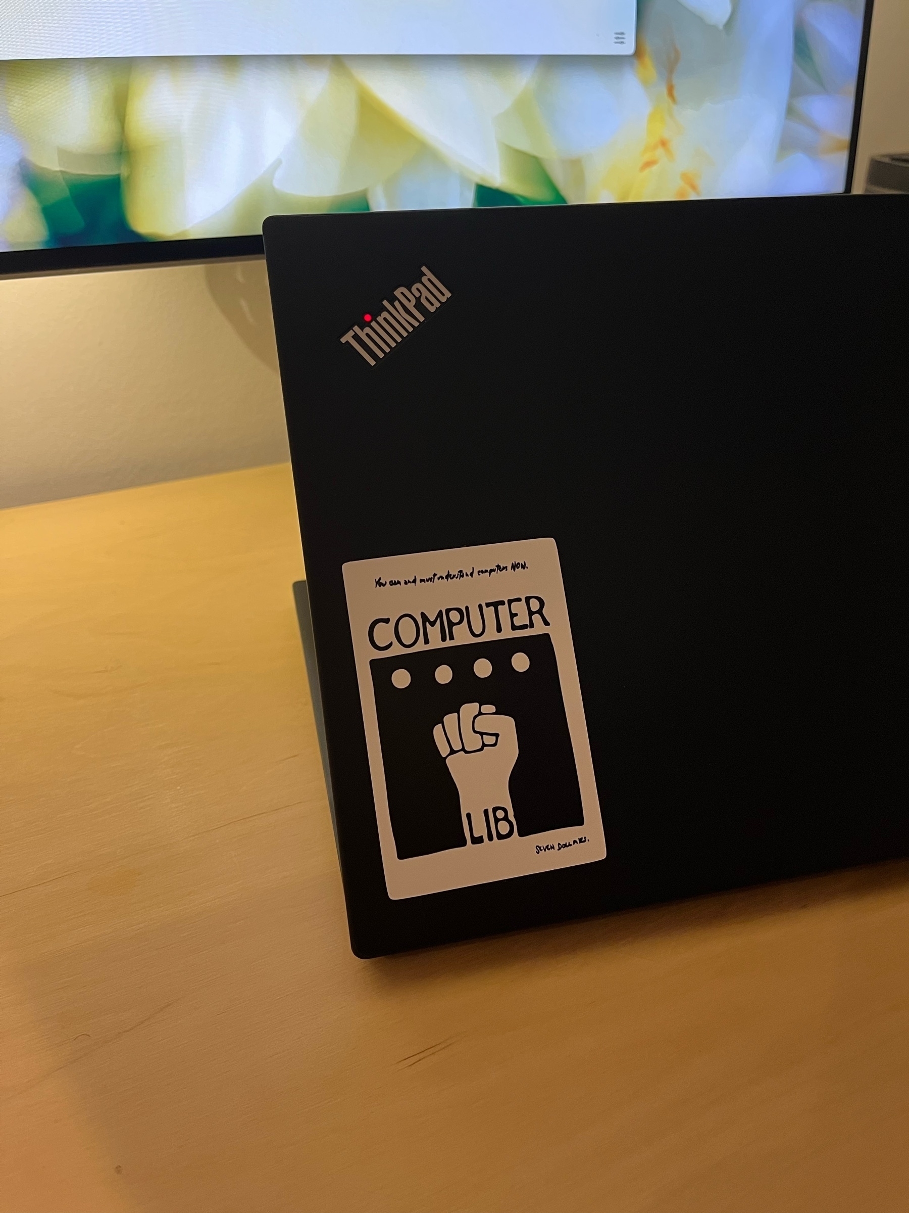 Laptop on a desk facing backwards with a sticker on the bottom left corner which contains the book cover, warm light.