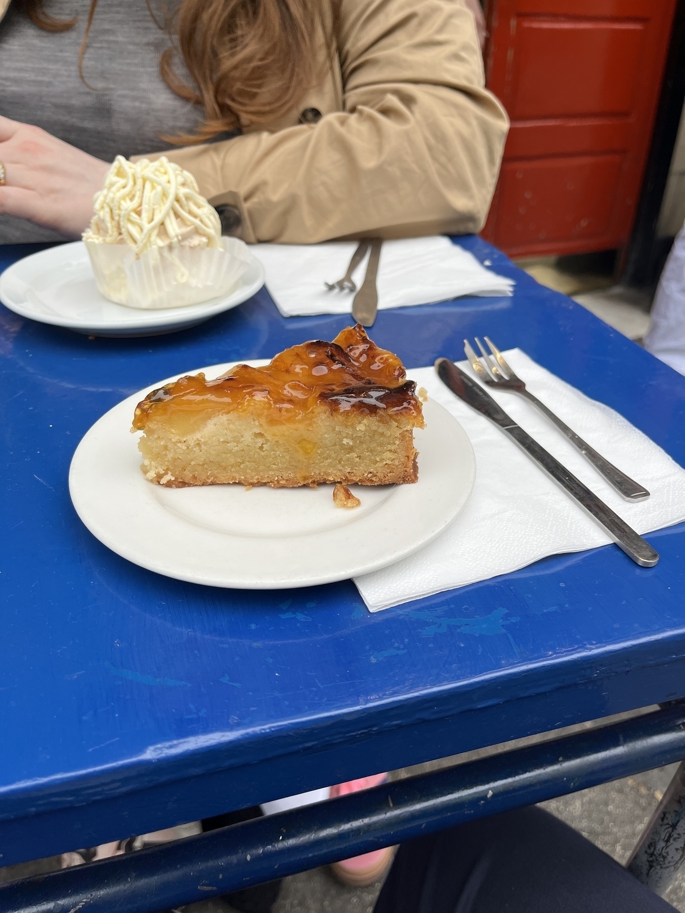 Triangle slice of Cake with almonds on top on a plate and blue bright table with knife and fork next to it.