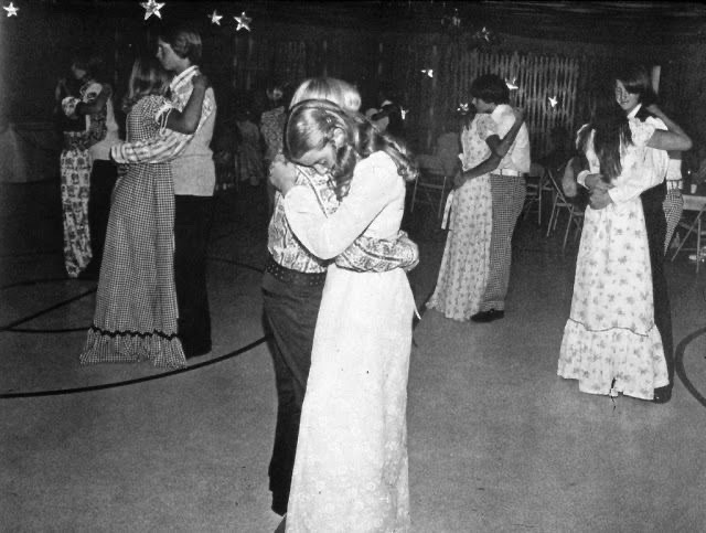 Black and white photo apparently from the mid-70s of teen-agers (they look to be about 15) slow dancing. The boys are wearing casual button down shirts and pants. The girls are wearing long dresses. The couples are pressed close together.