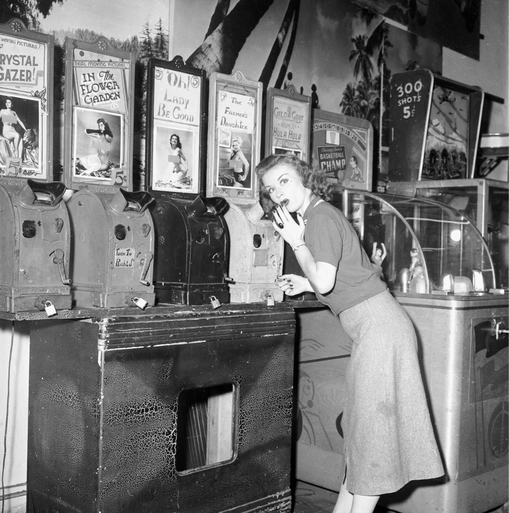 A black-and-white photo, I guess from the 1930s, of a woman in a dress and sweater standing in front of a row of kinetoscopes (those old timey one-person movie machines that you peered into). She’s bent slightly in front of a machine with the title “The Farmer’s Daughter,” and has her hand to her mouth, pretending to look shocked.