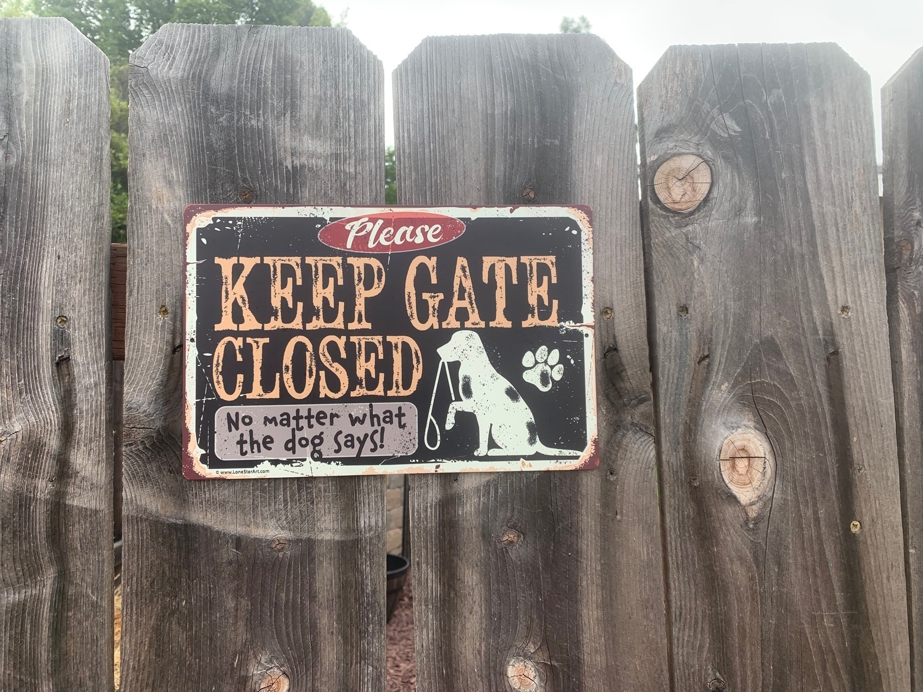 Weathered sign on wooden gate. Text: “KEEP GATE CLOSED: No matter what the dog says!”