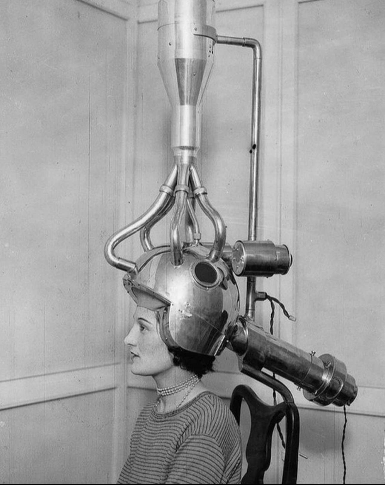A black and white vintage photo of a woman sitting with an elaborate hair dryer machine (?) fitted over her head. The machine has multiple tubes and a large metal hood.