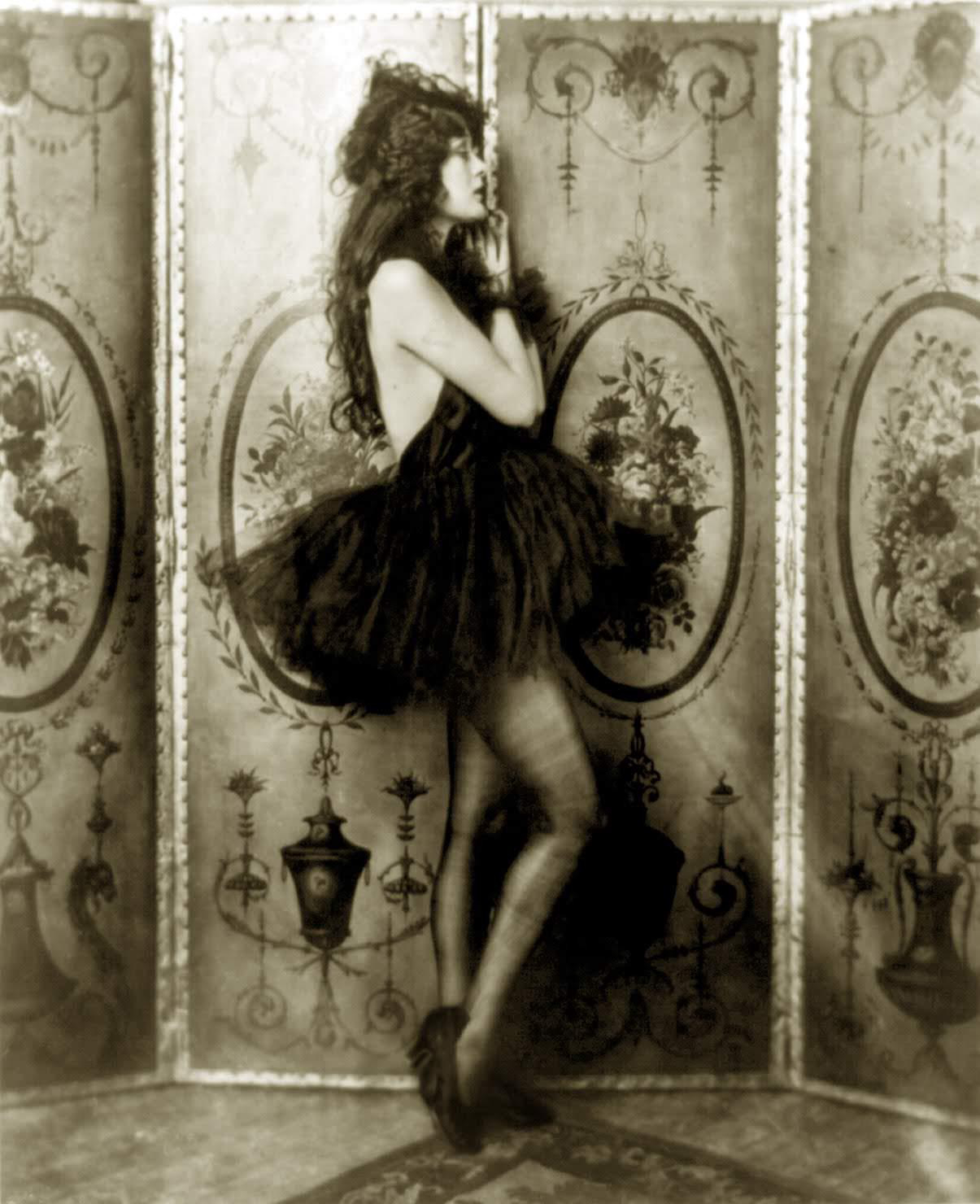Vintage sepia-toned photograph of a pretty woman in a ruffled tutu and headdress, posing with one leg lifted, against a decorative folding screen.