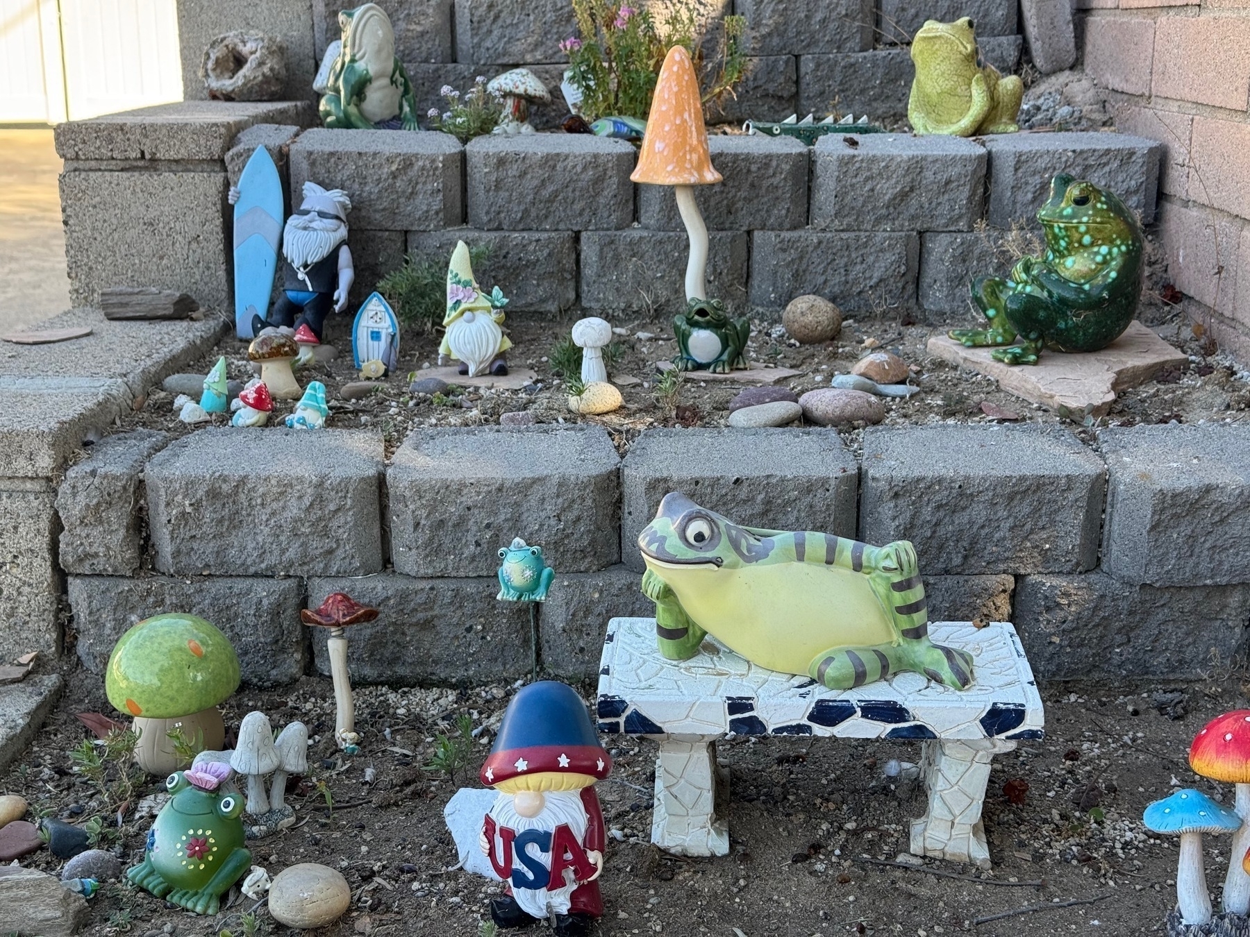 Two of the terraced steps, with figurines of a frog reclining on a park bench, little red-white-and-blue patriotic garden gnome, surfin' Santa, and more