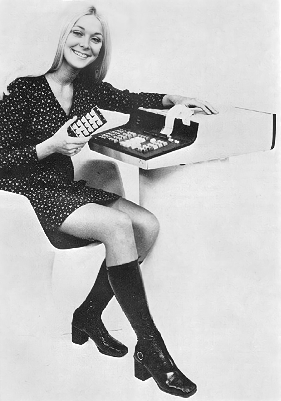 Black-and-white photo from about 1970 of a smiling long-haired slender blonde woman in a short dress and go-go boots sitting with a retro computer keyboard and cassette tapes.