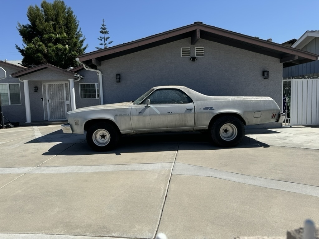 Auto-generated description: A vintage beige car is parked on the driveway of a suburban house with a gray exterior.