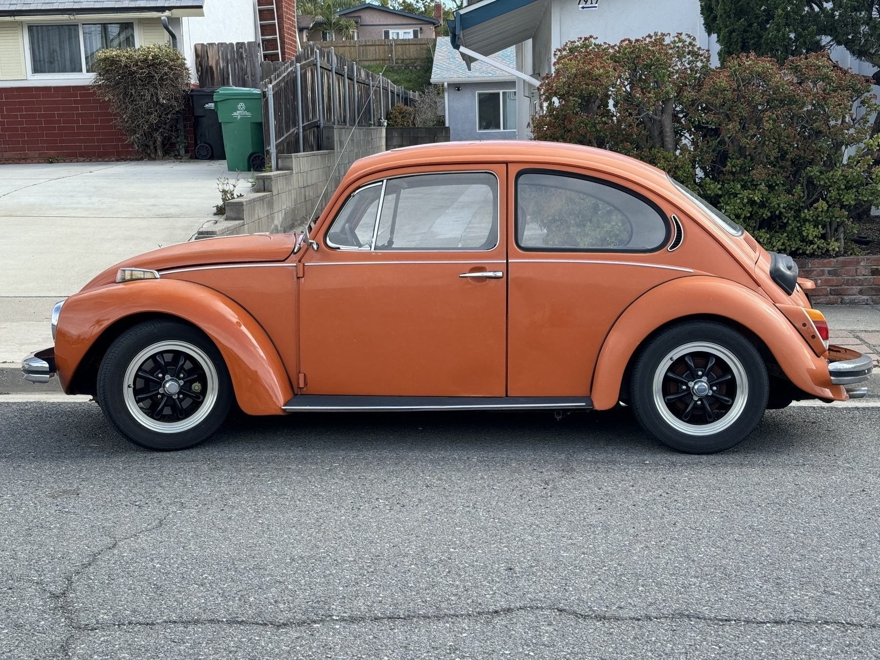 Orange vintage Volkswagen Beetle, viewed side-on, nicely centered in the photo, parked in front of and between two suburban houses.