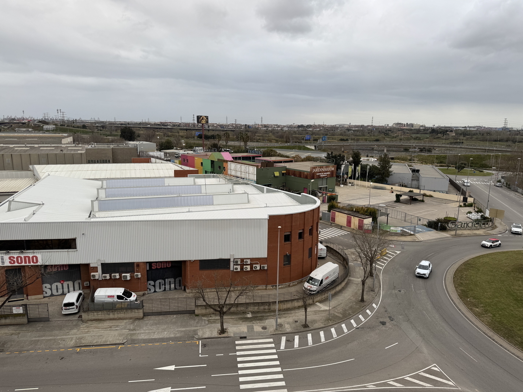 A view from above shows a curving road beside industrial and commercial buildings, with a cloudy sky and open landscape in the background.