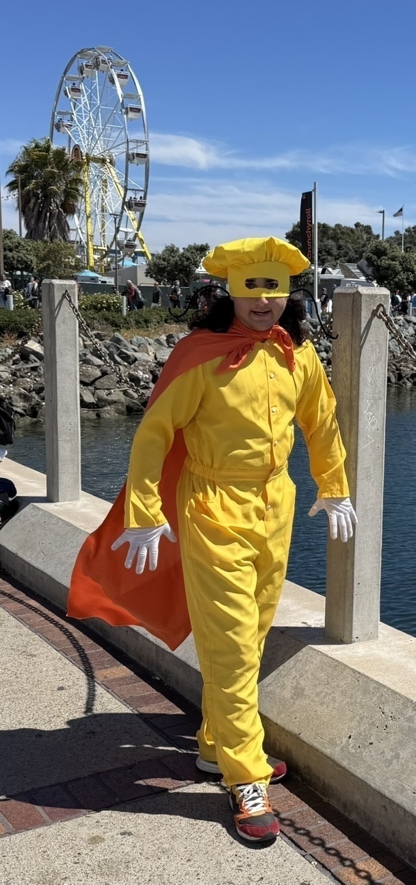 Auto-generated description: A person in a bright yellow costume with a red cape and white gloves is walking along a waterfront, with a ferris wheel visible in the background.