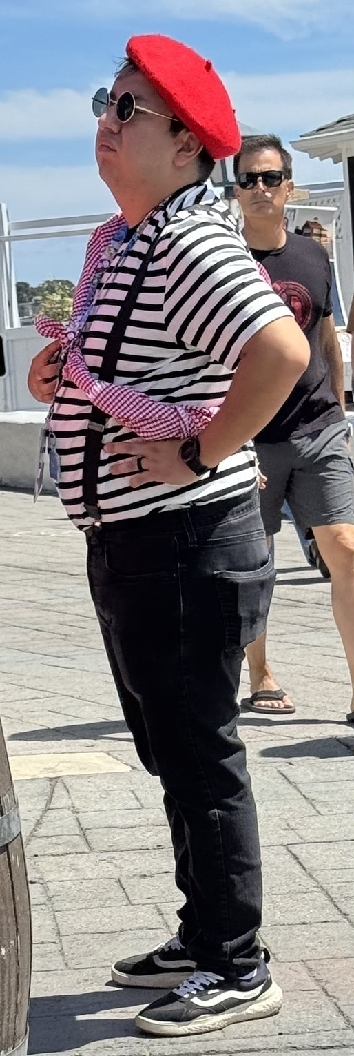 Auto-generated description: A person wearing a red beret, round sunglasses, striped shirt, and suspenders stands outdoors with a hand on their hip.