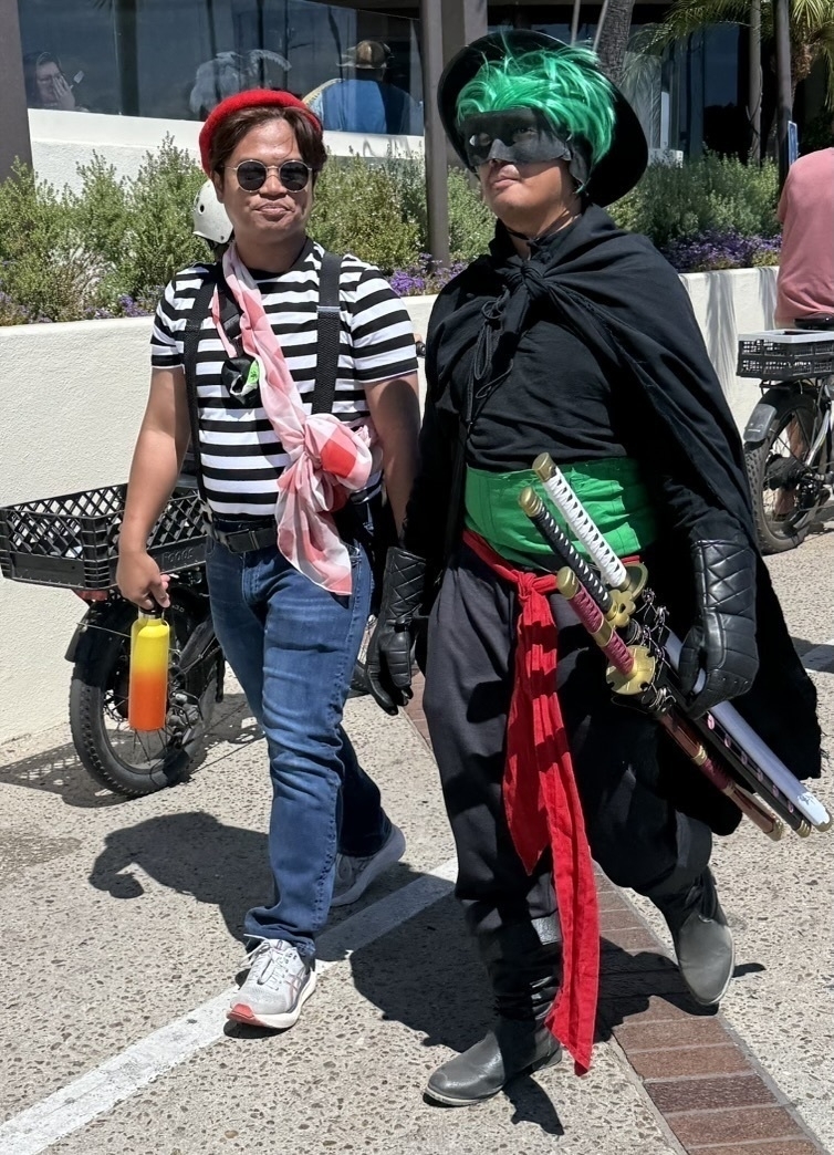 Auto-generated description: Two people are walking, one dressed casually and the other wearing a costume with green accents, carrying swords.