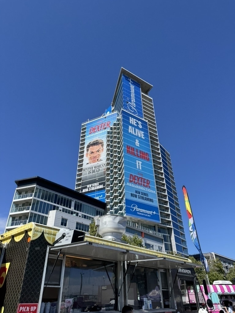 Auto-generated description: A tall building features large advertisements, primarily for Dexter, against a clear blue sky, with food stalls in the foreground.
