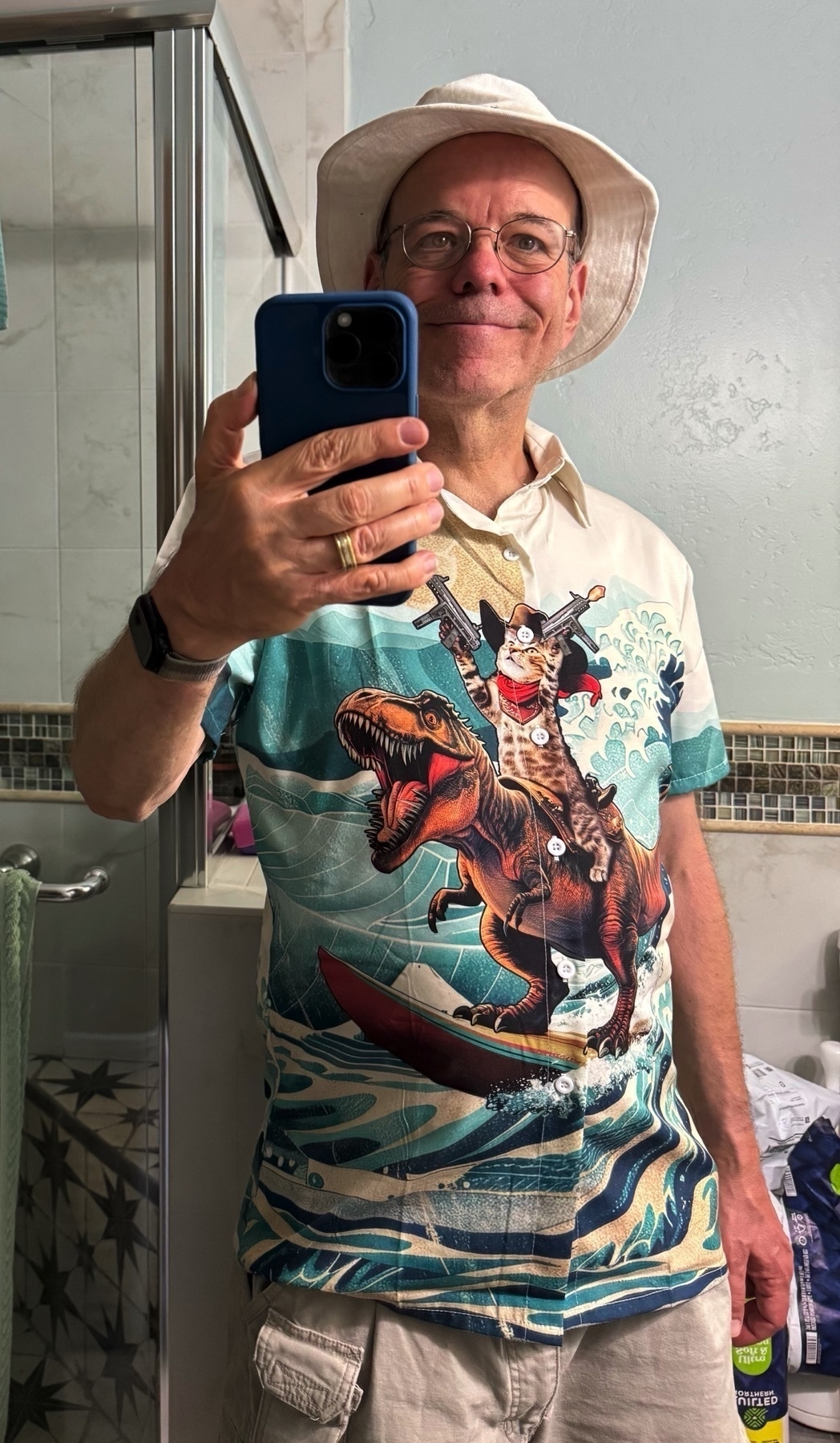 Auto-generated description: A man wearing a hat takes a selfie in a bathroom mirror, showcasing his colorful shirt featuring a surfing dinosaur.