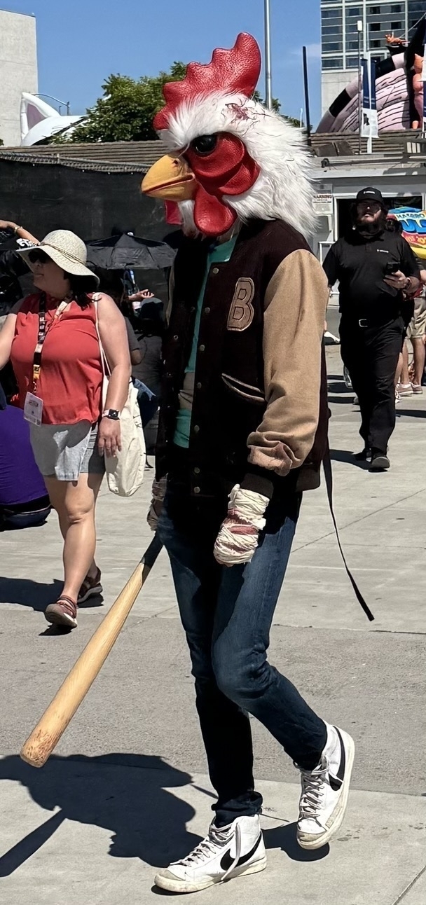 Auto-generated description: A person wearing a large rooster head mask and holding a baseball bat walks down a sunny street.