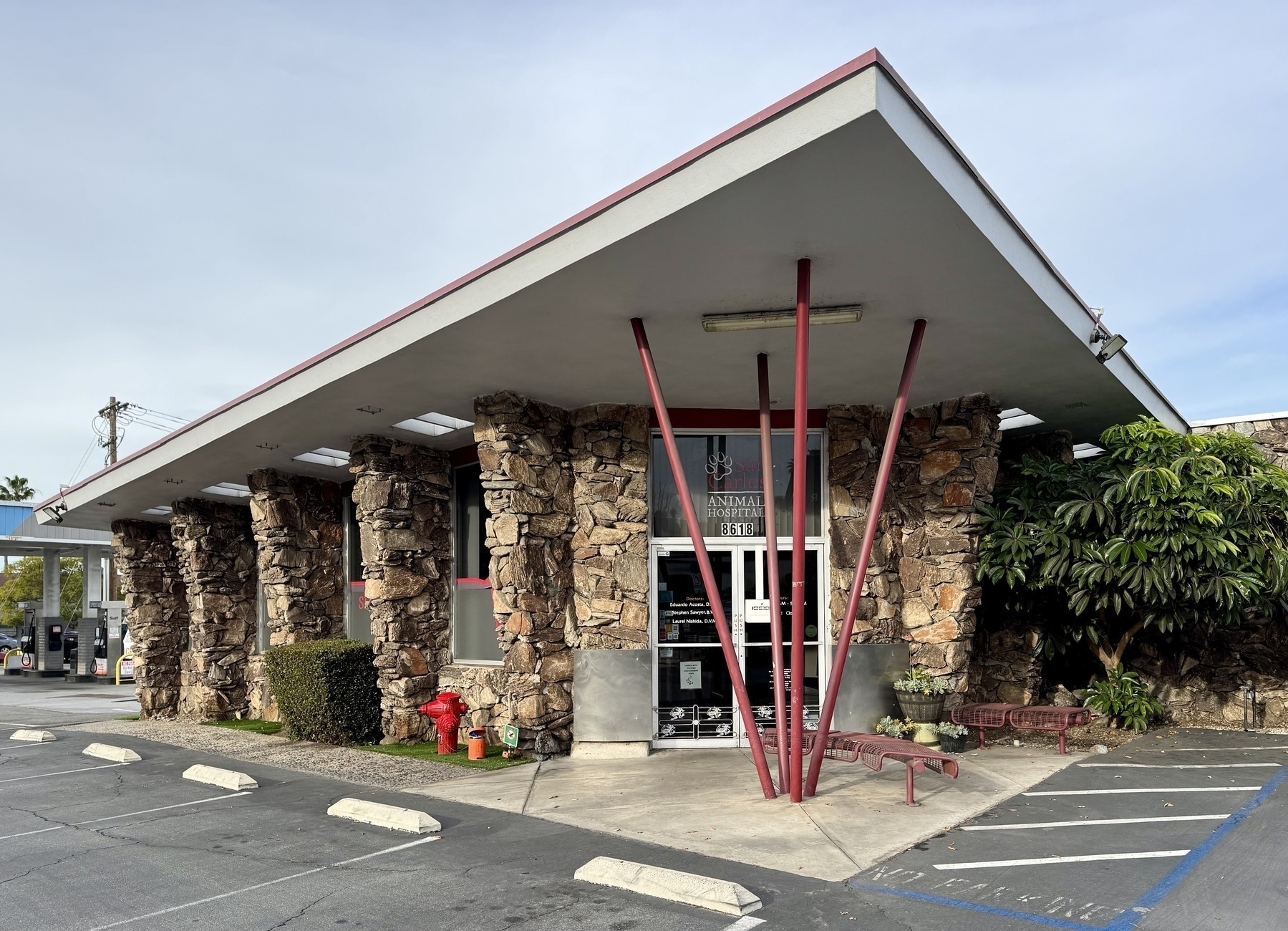 A mid-century modern building features a distinct slanted roof, stone facade, and red support beams, situated next to a parking area.