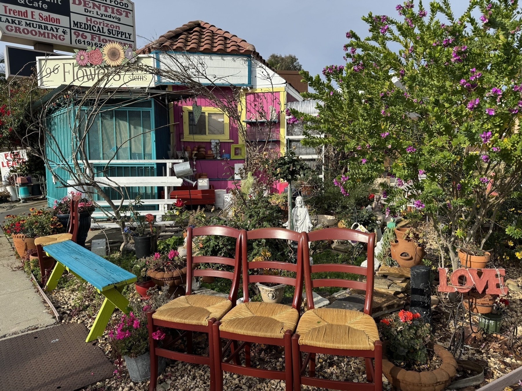 A colorful garden setting features a small flower shop, vibrant plants, three red chairs, and various decorative items including a LOVE sign.