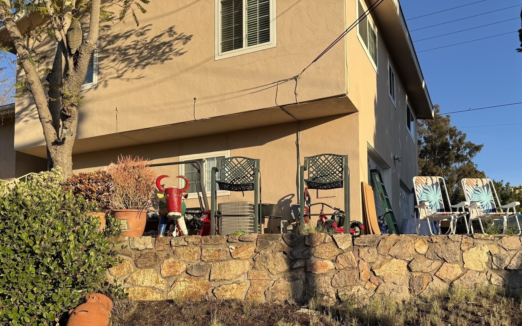 Stone wall borders a garden with chairs and potted plants and a red metal bull sculpture in front of a two-story house.