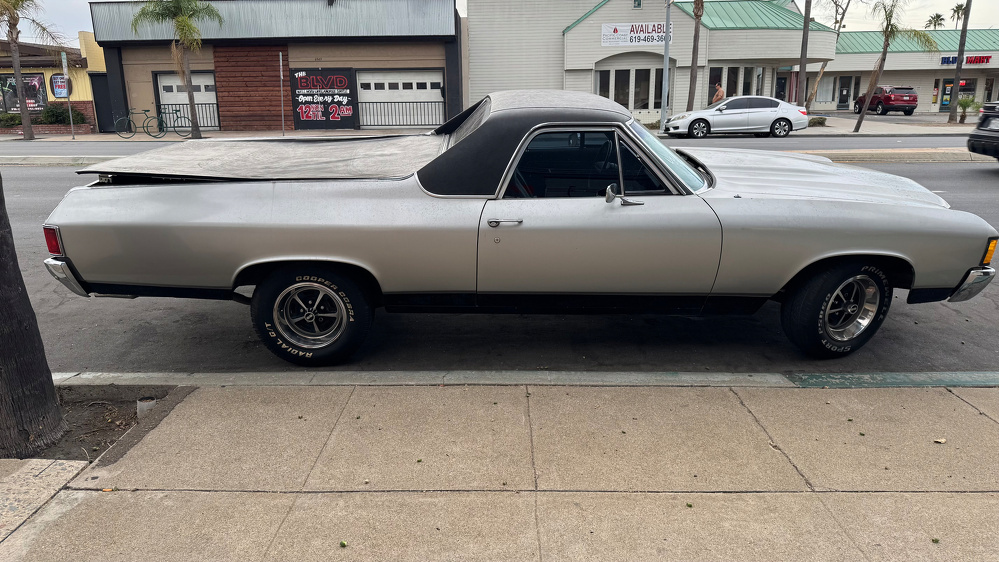 A silver classic car with a black soft top and covered bed is parked on the street in front of a row of shops.