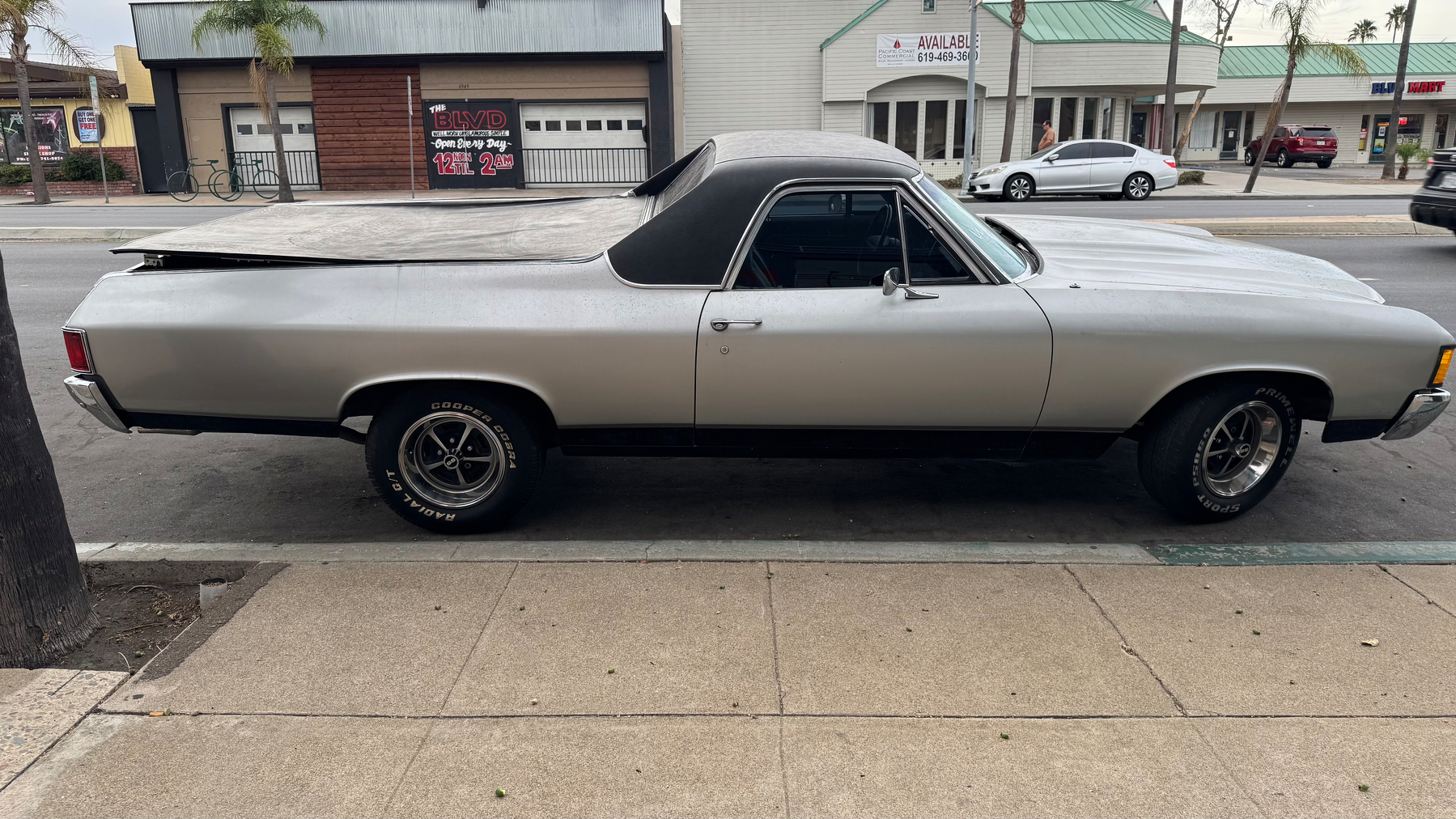 A silver classic car with a black soft top and covered bed is parked on the street in front of a row of shops.