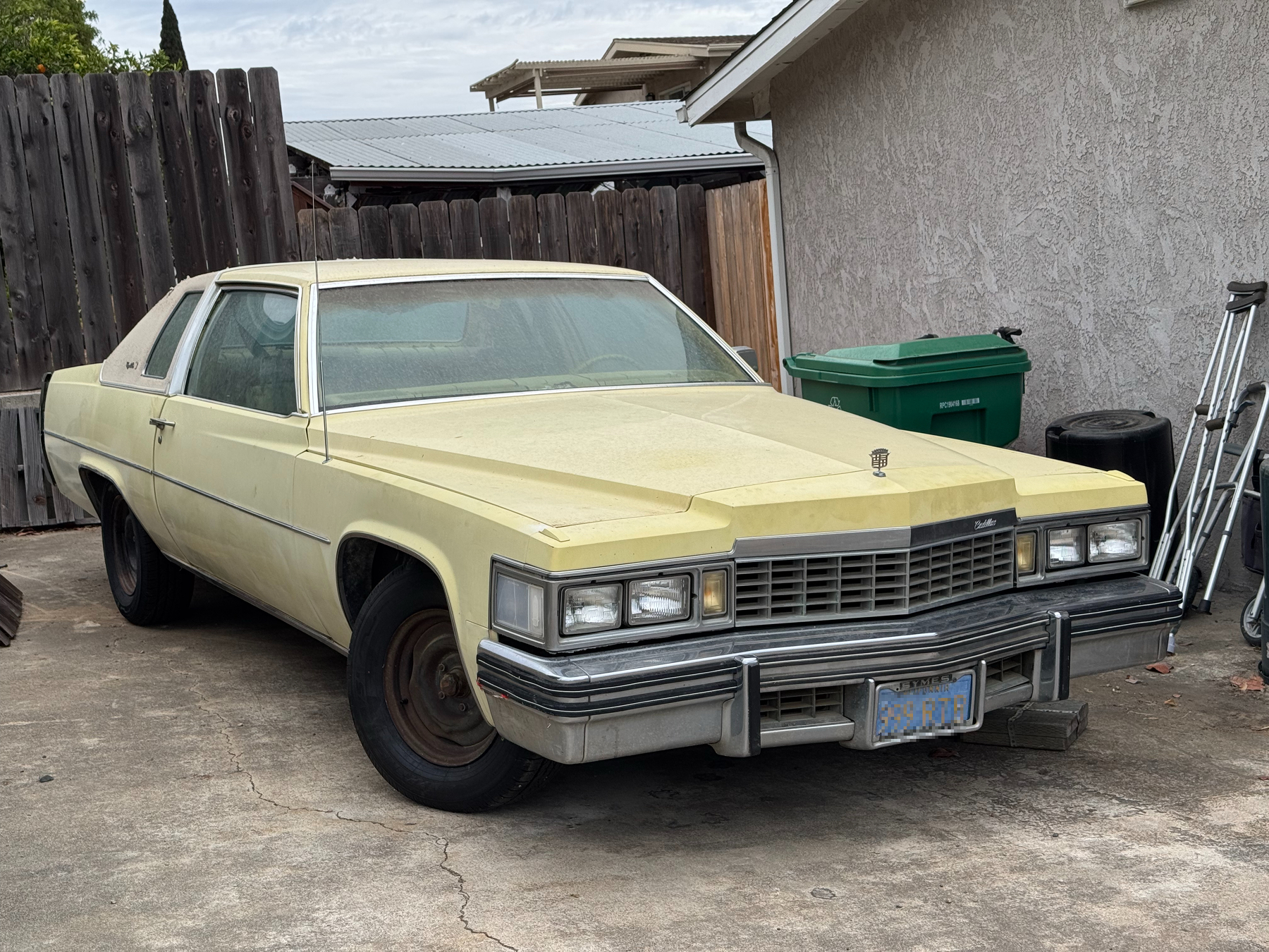 Dusty pale yellow late-1970s Cadillac Coupe de Ville with a landau roof and bare steel wheels, parked on a cracked driveway beside a wooden fence