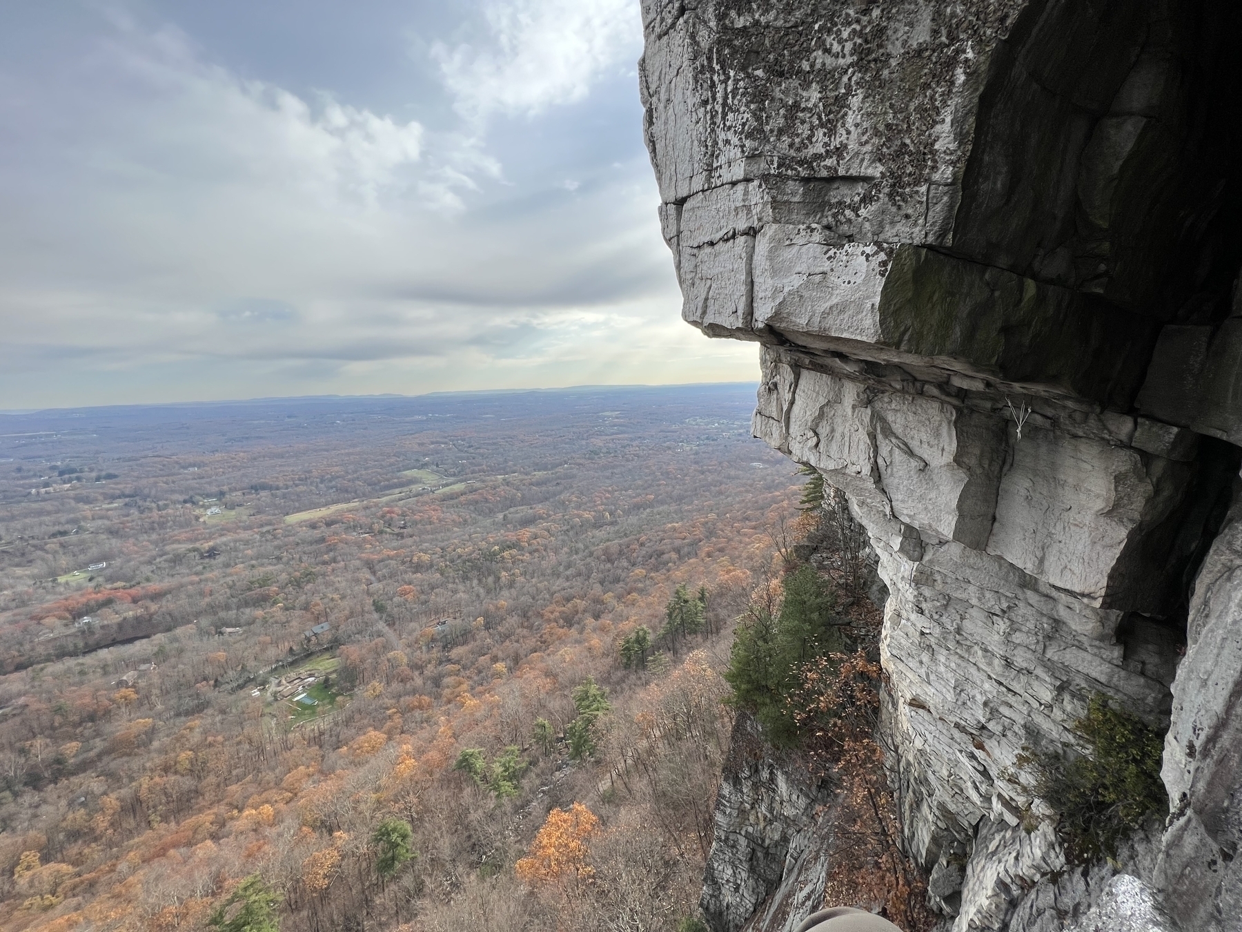 Looking down at the rest of the Gunks cliff where there’s another climber and trees far below in the background.