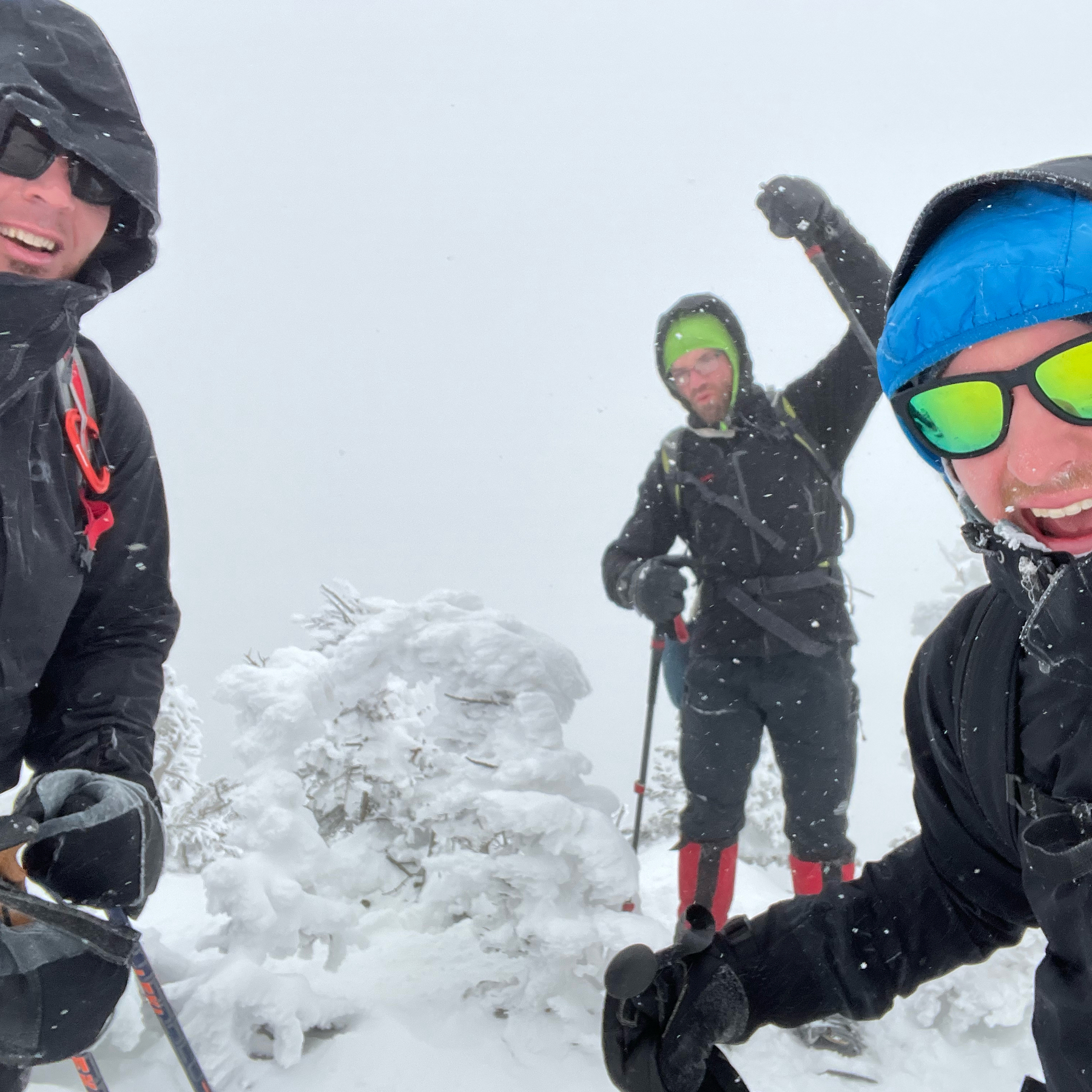 Three guys, dressed in winter gear and with snow and ice frozen on their faces, in celebration at the summit of a mountain.