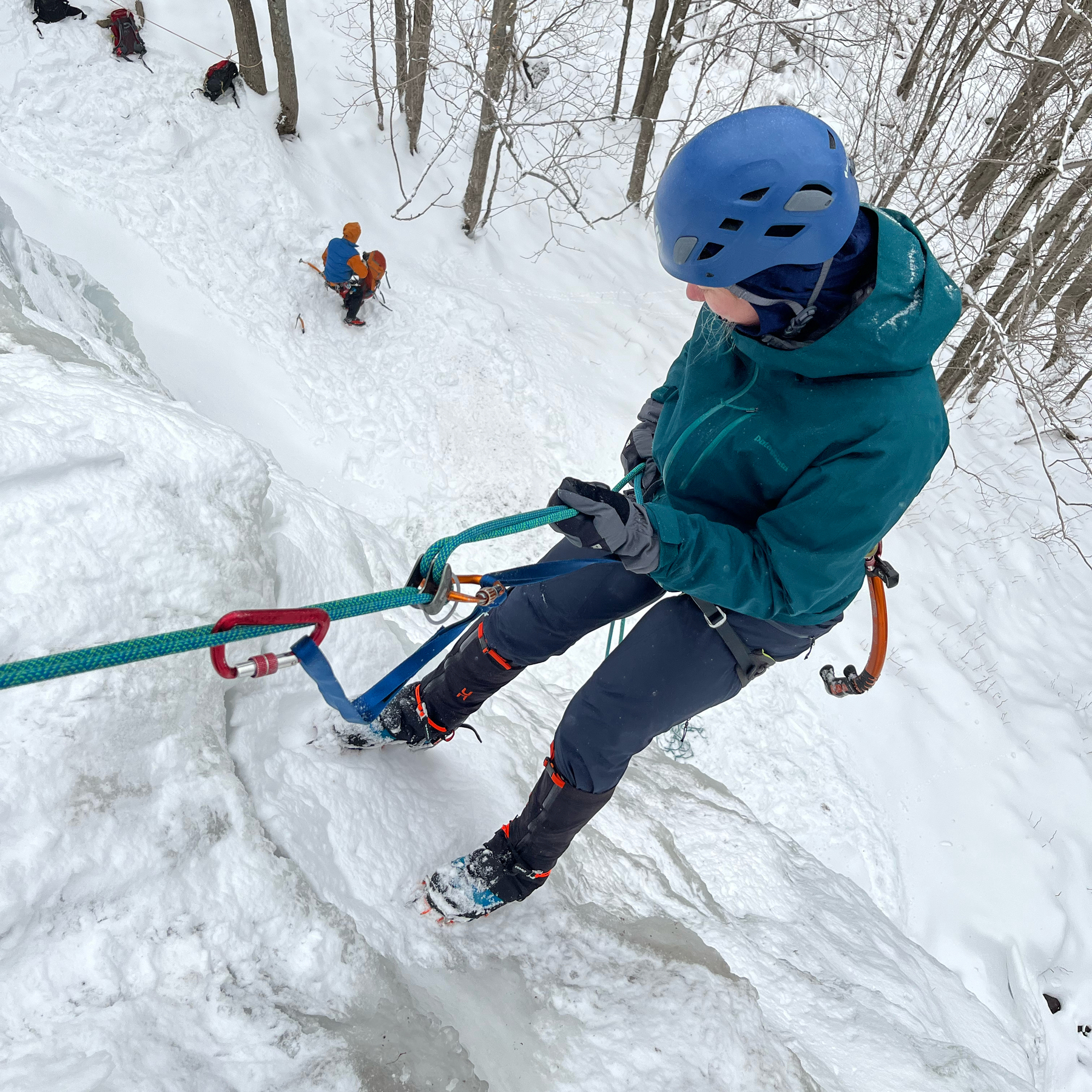 A girl in a blue winter coat rappels down an ice clif. Taken from above.