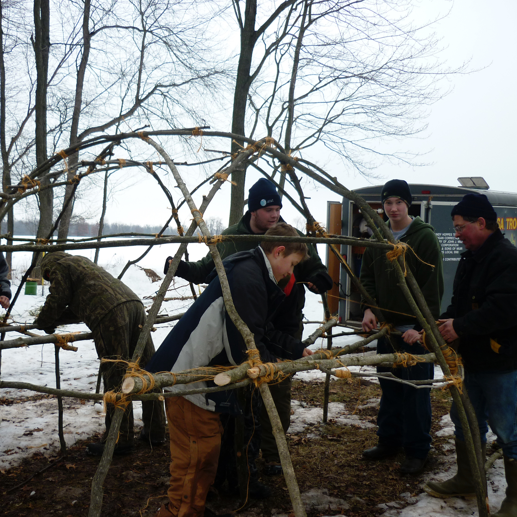A wigwam made of sticks under construction by Boy Scouts in the winter.