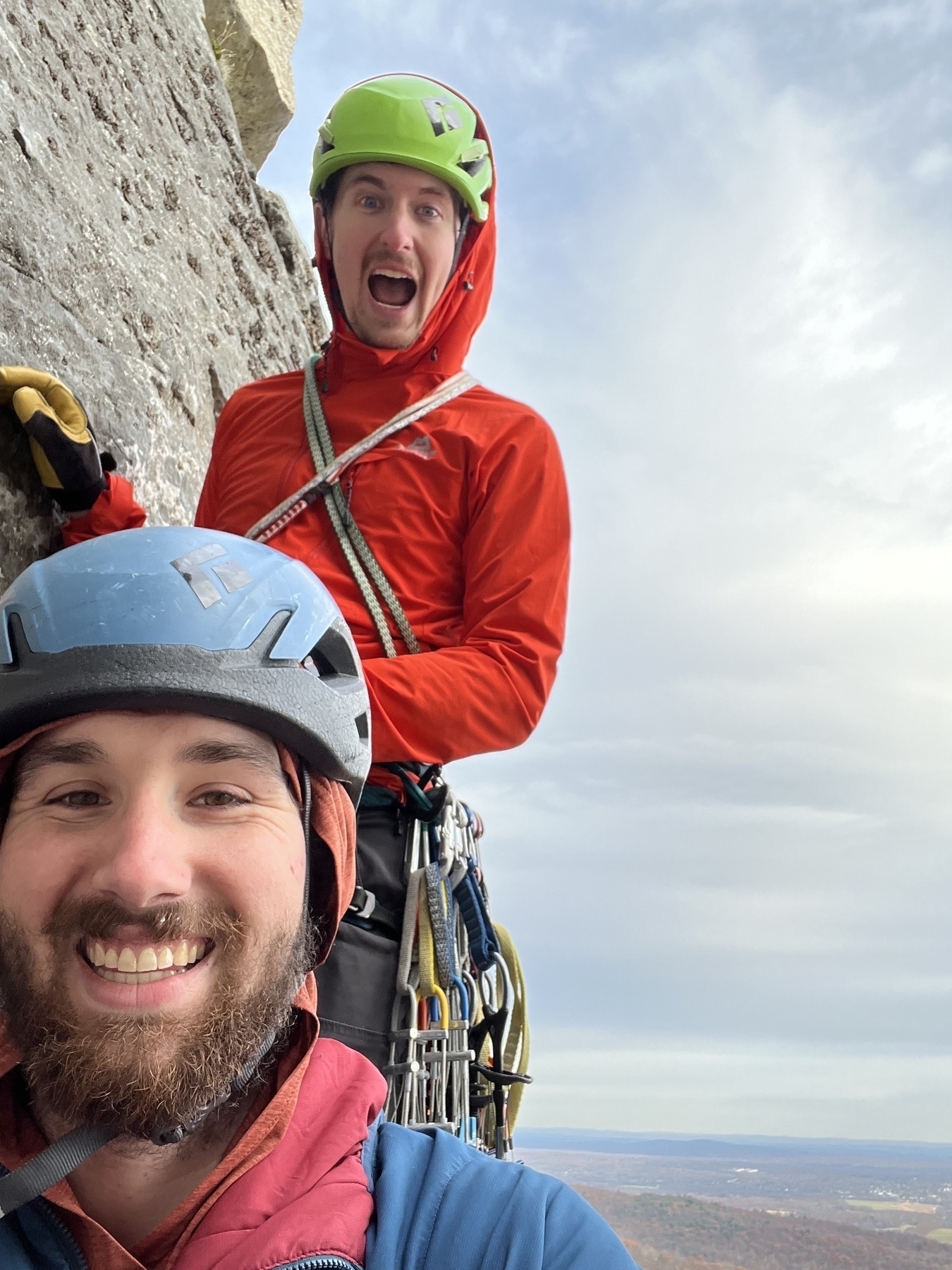 Two men in climbing gear smiling at the camera with a lard rock wall to their right.