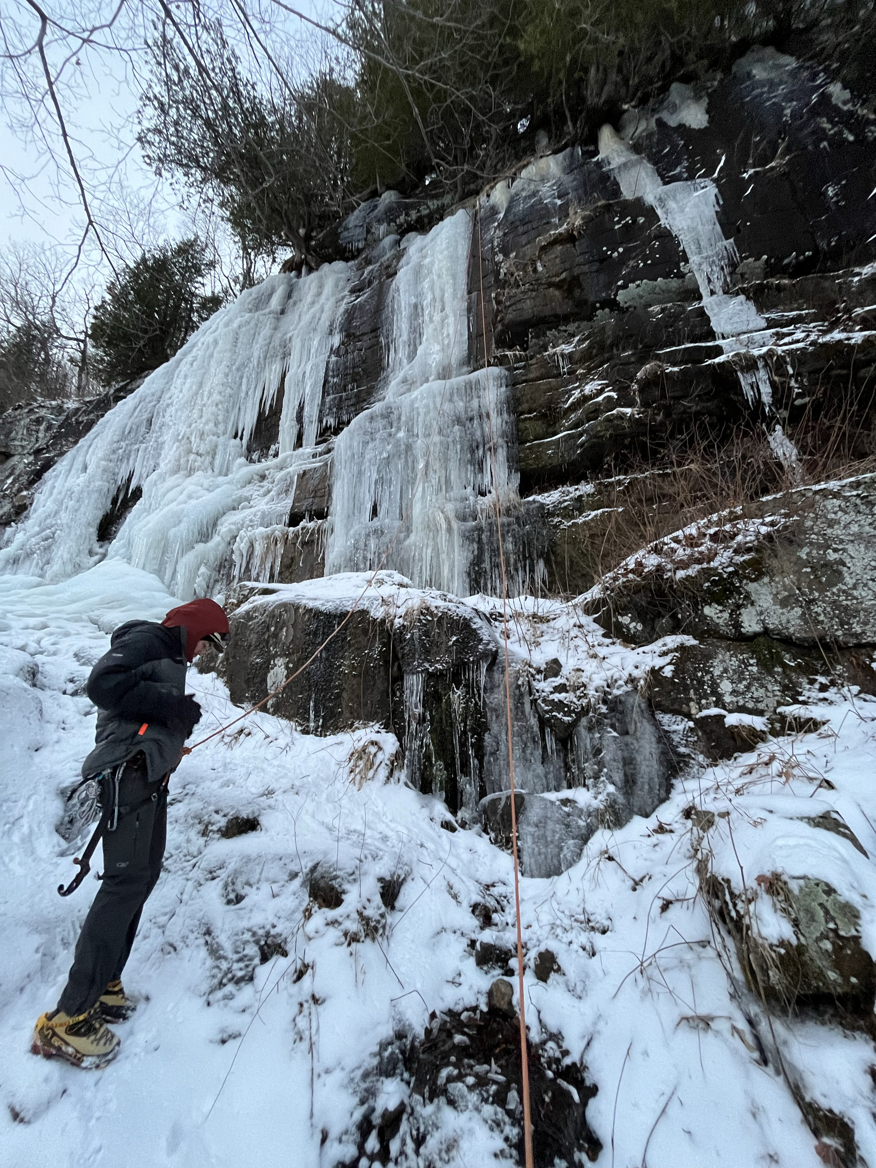 Man standing below a frozen waterfall, dressed in ice climbing gear.