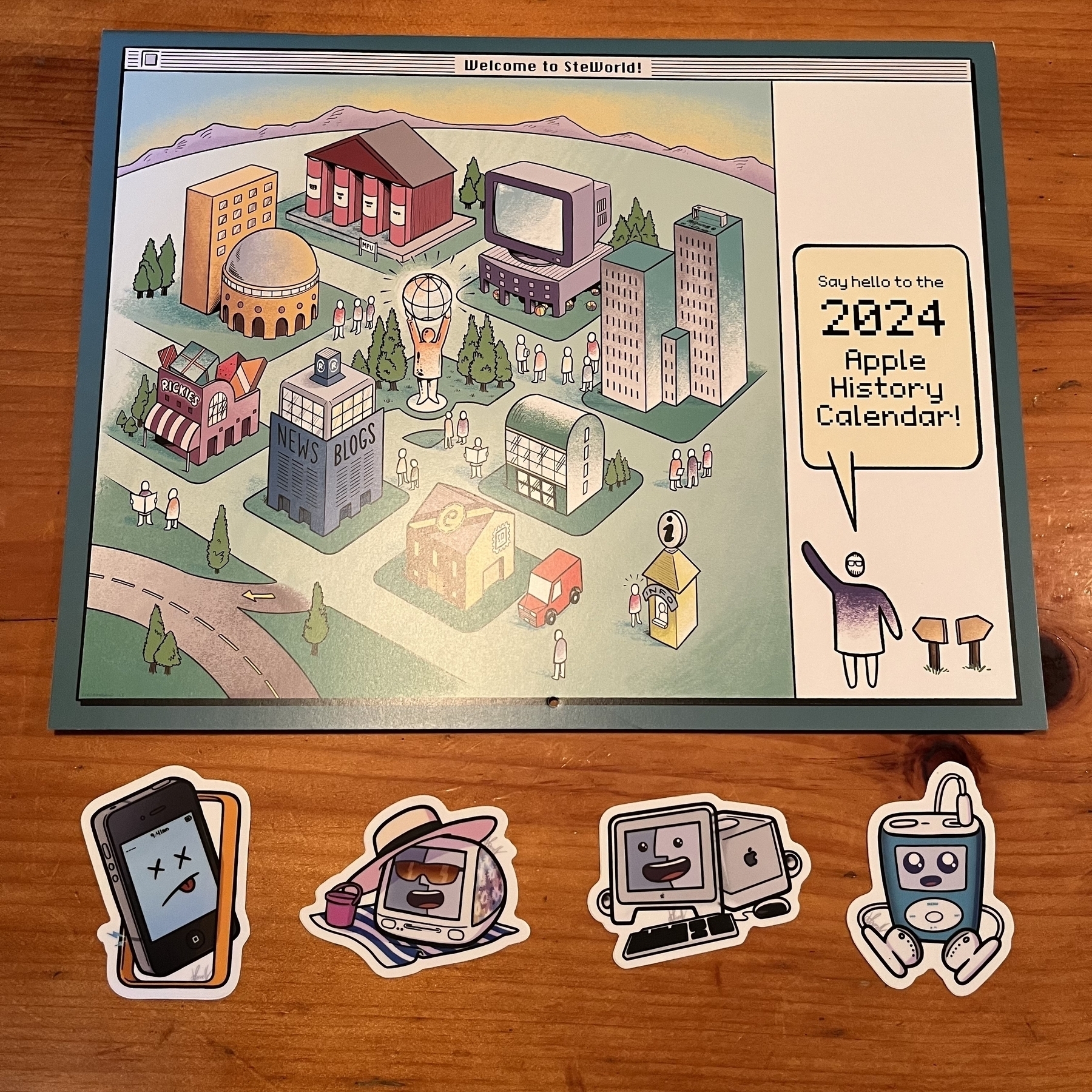 The cover of the 2024 Apple history calendar that shows the bird side view of a cartoon-like city, and four stickers themed around the iPhone, iMac, and iPod.