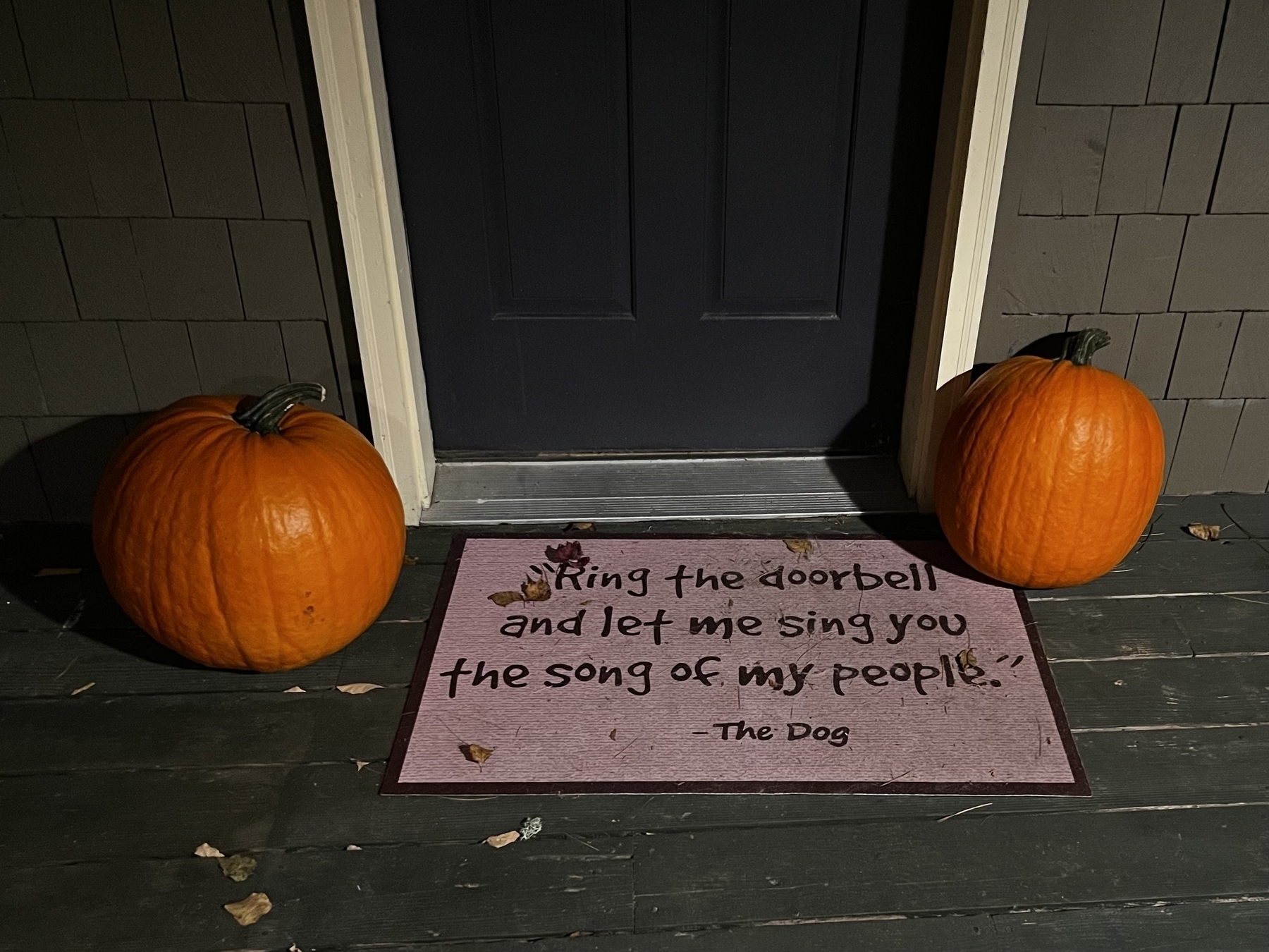 Two uncarved pumpkins on either side of a closed door and a doormat that that reads, &ldquo;ring the doorbell and let me sing you the song of my people&rdquo; with a dog paw print.
