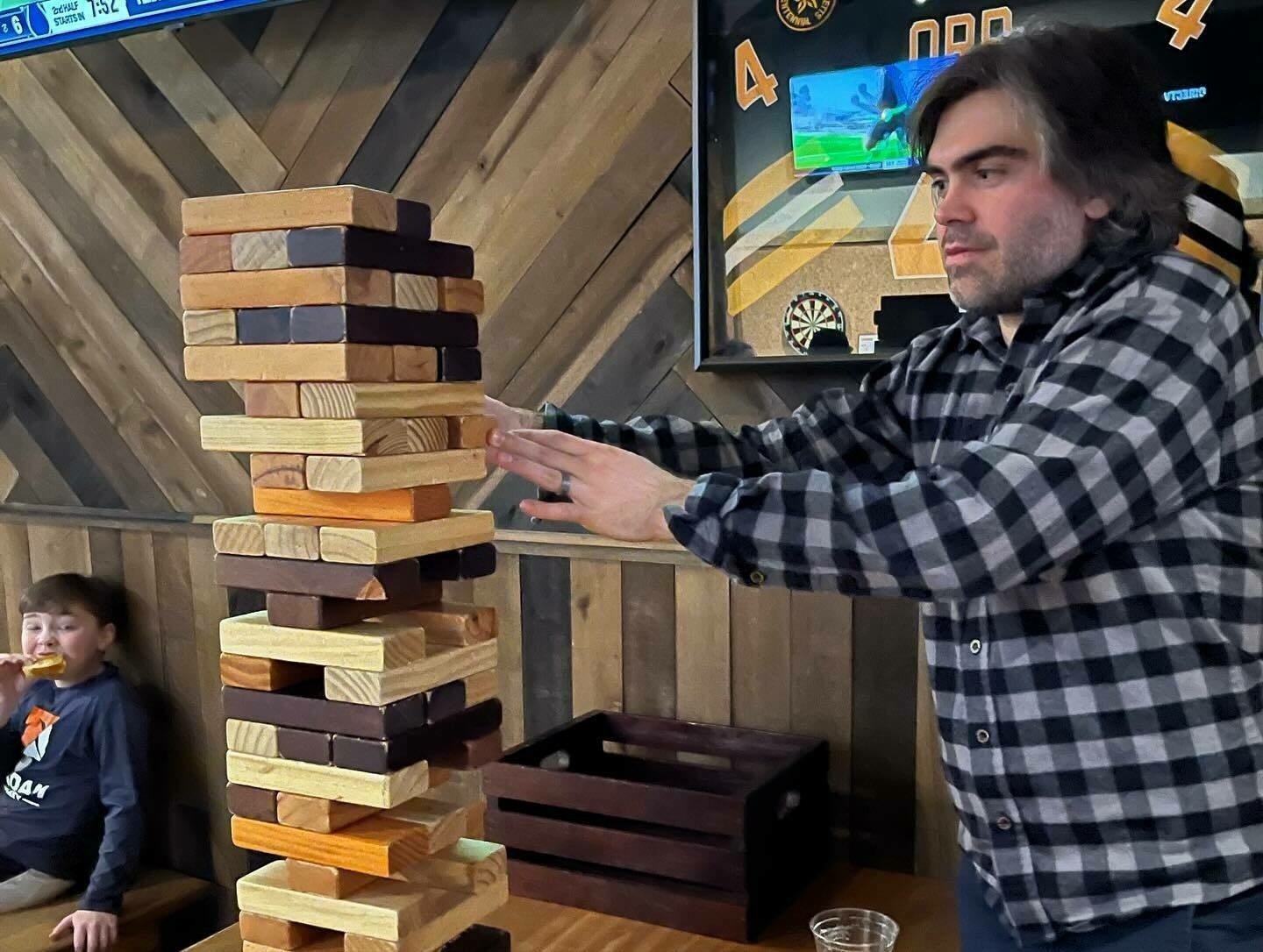 A man is carefully pulling a wooden block from a large, stacked Jenga tower in a casual indoor setting while a child, eating, watches in the background.