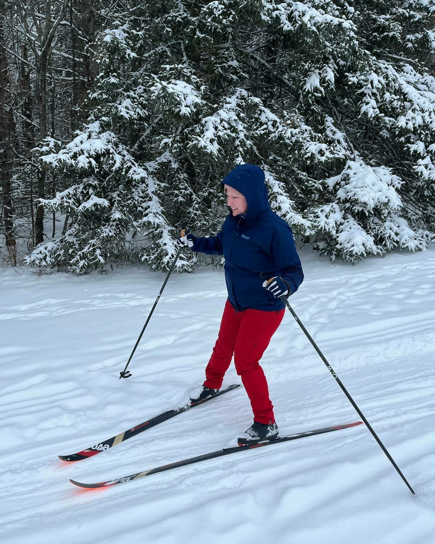 A person is cross-country skiing on snow-covered ground, flanked by snow-laden evergreen trees. They are dressed in winter gear, including a blue jacket and red pants.