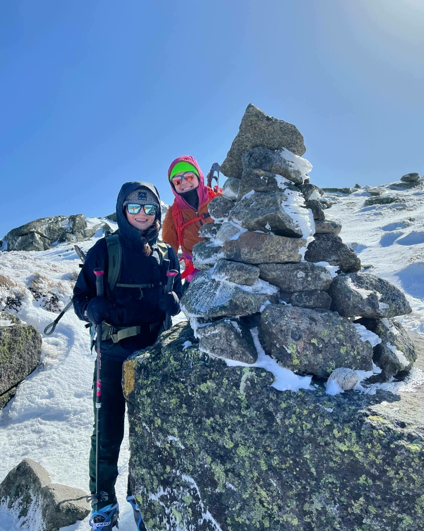 Two individuals are posing behind a cairn on a sunny, snow-covered mountain. They are dressed in vibrant cold-weather gear, with hiking equipment.