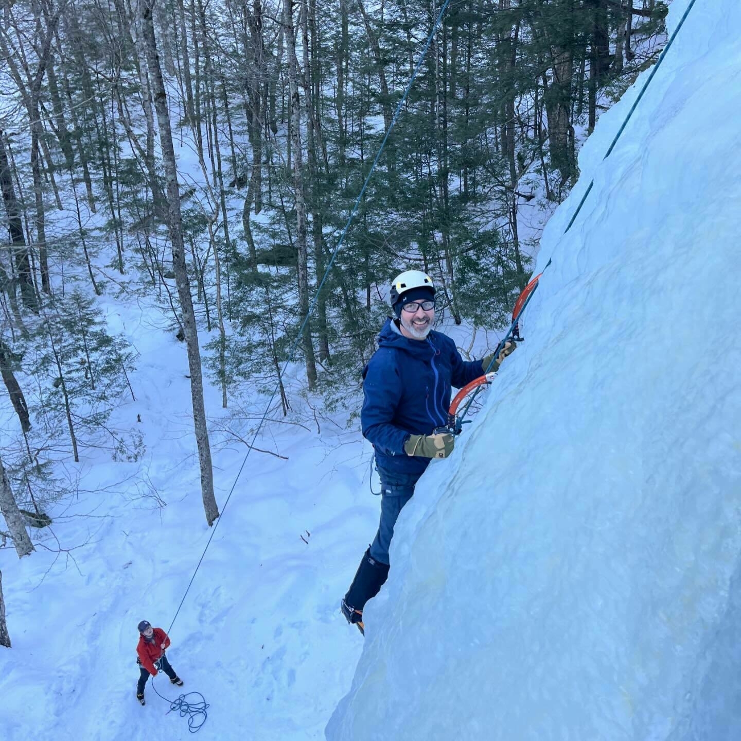 A person is ice climbing a frozen slope while another person stands below on snow-covered ground, surrounded by a forest of bare trees. The climber is smiling and secured by ropes.