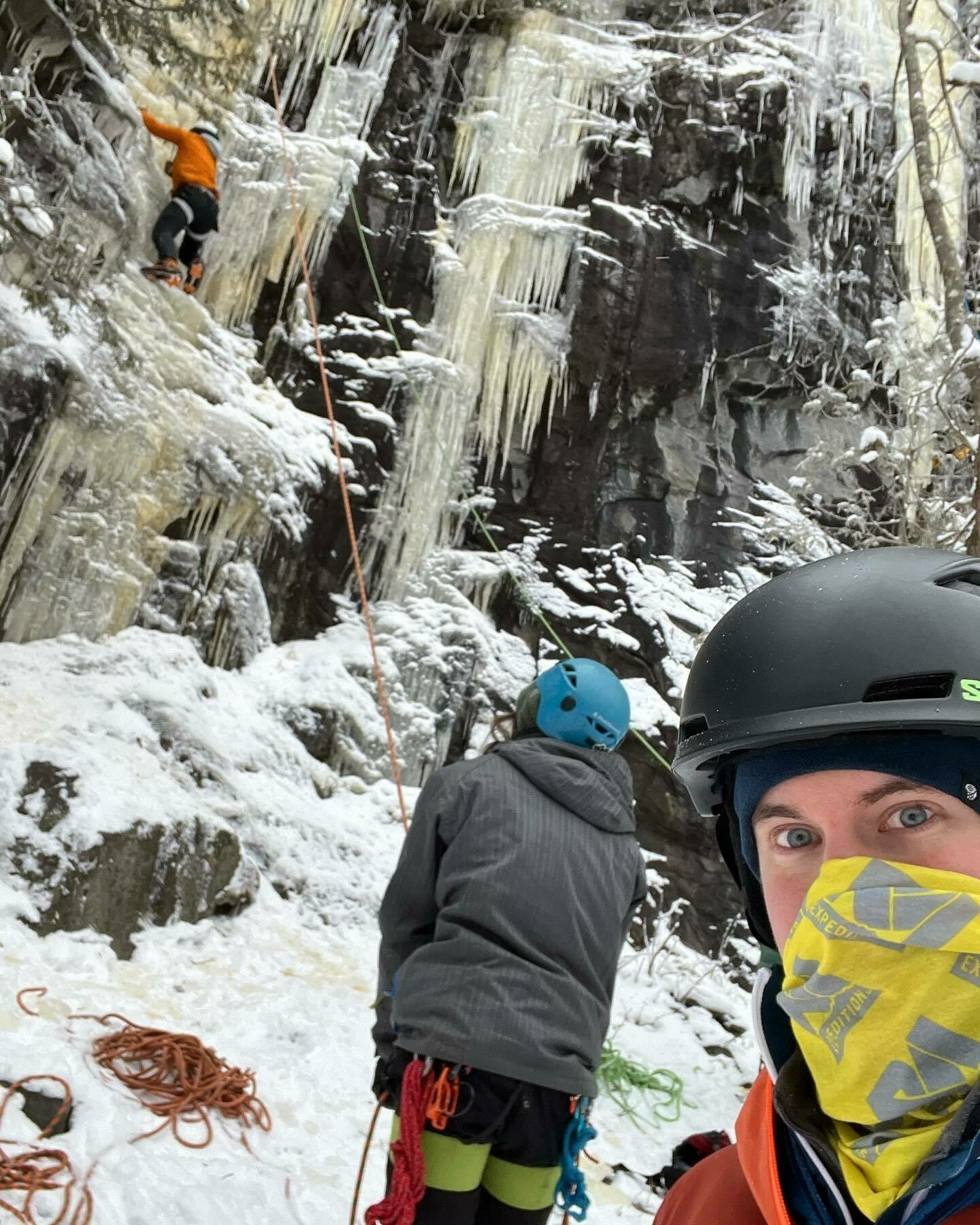 A climber ascends a frozen waterfall, secured by ropes, while two others watch. They are equipped with climbing gear in a snowy, icy environment.