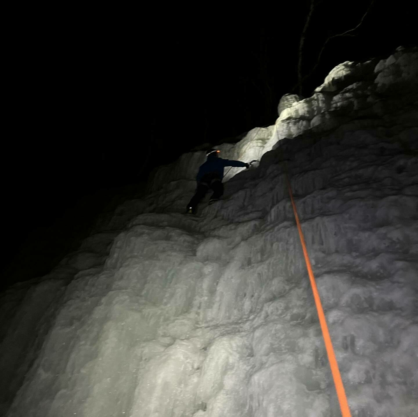 A person is ice climbing a steep frozen wall at night, secured by an orange rope, with darkness surrounding them.