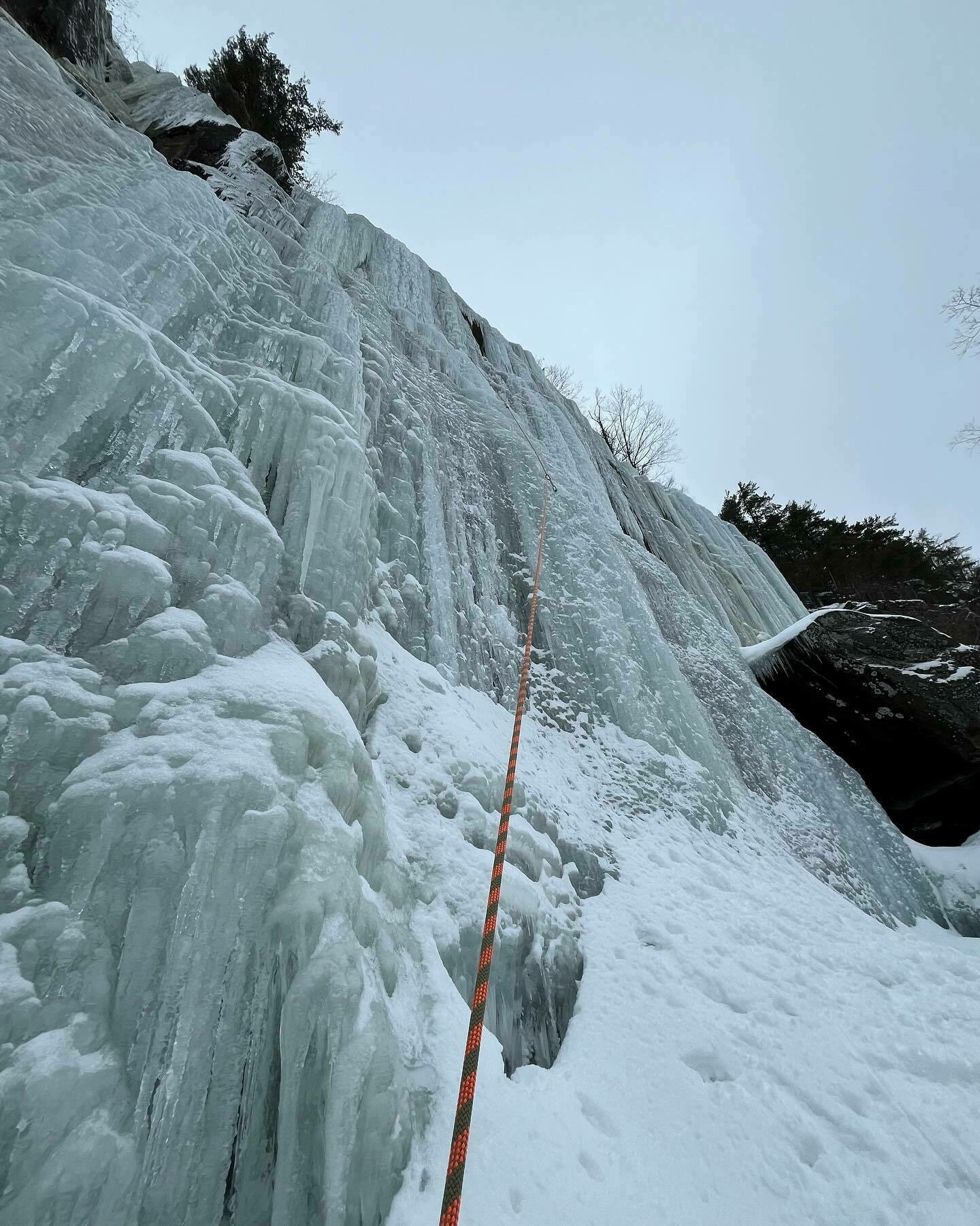 An orange climbing rope stretches up a steep, frozen waterfall with intricate ice formations, set against a forested backdrop.