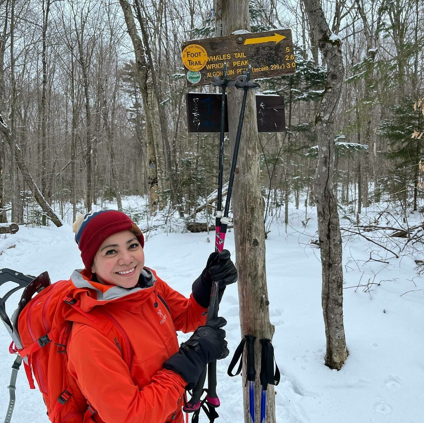 A smiling person holding hiking poles stands next to a trail sign in a snowy forest. The sign reads &ldquo;FOOT TRAIL → WHALES TAIL 5 WRIGHT PEAK 2.6 ALGONQUIN PEAK 2994'↑3.0.&quot;