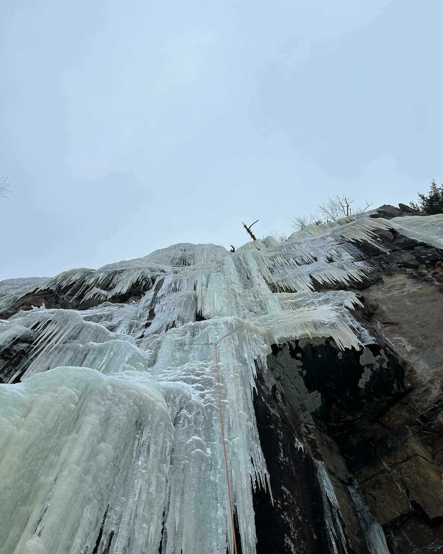 A person ice climbing a frozen waterfall with climbing ropes, against an overcast sky.