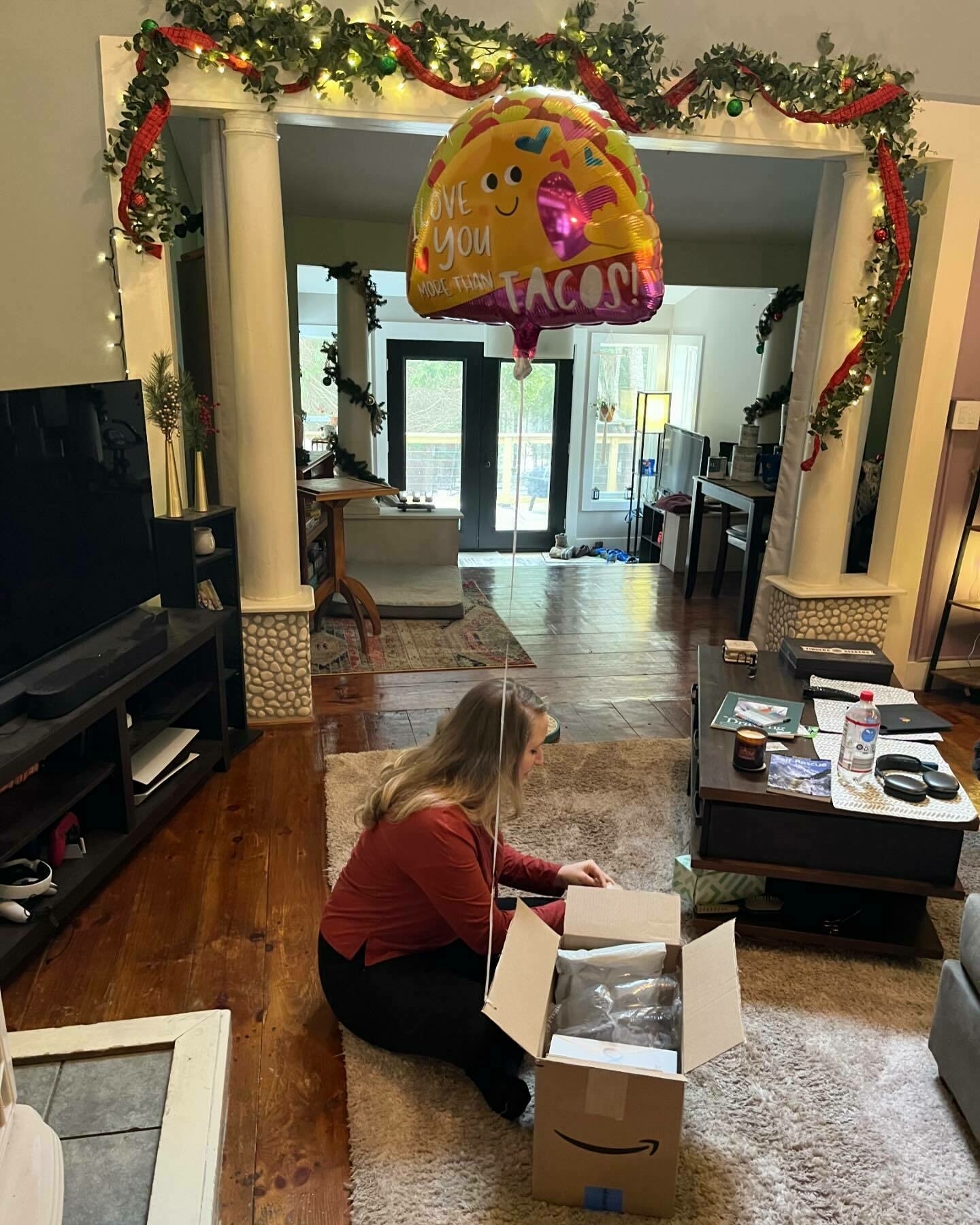 A person sits on the floor unpacking a box in a decorated living room. A balloon overhead reads &ldquo;I Love You More Than Tacos!&quot;
