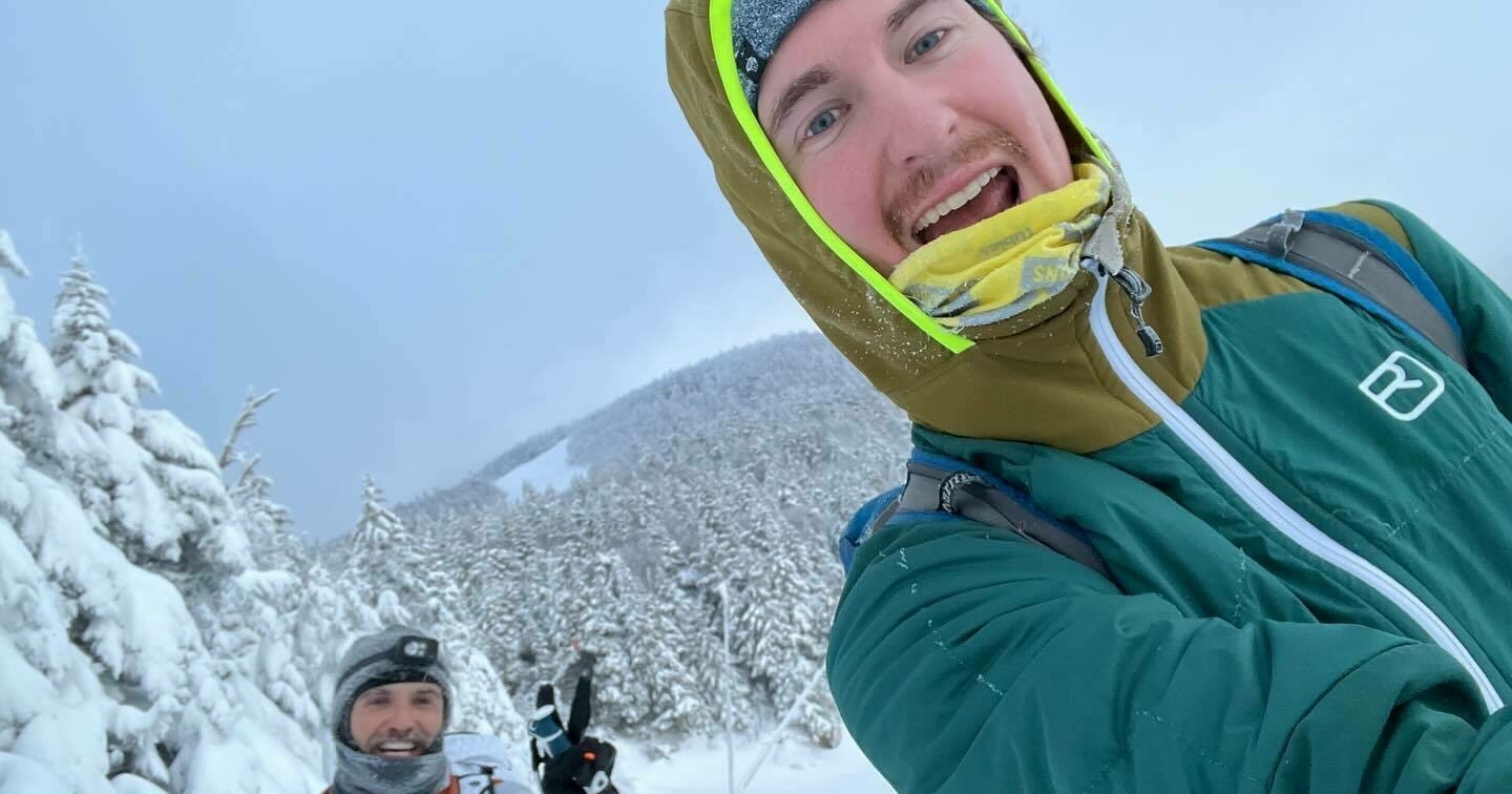 Two people are taking a selfie in a snow-covered landscape; one in the foreground smiles broadly, and another slightly behind wears a headlamp. Trees and a mountain form the backdrop.