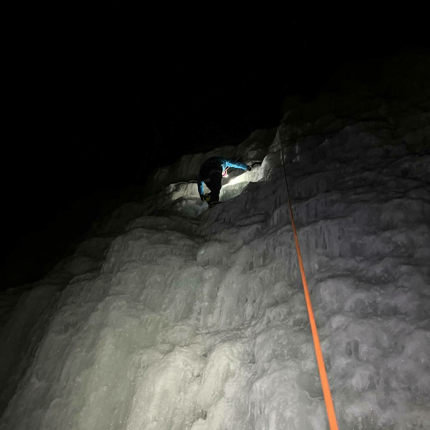 A person is ice climbing at night, illuminated by a headlamp, with an orange rope extending downward in a dark environment.
