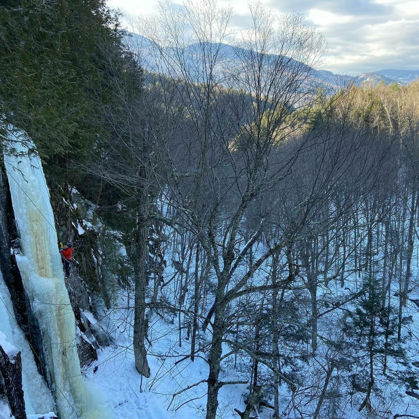 A person is ice climbing on a frozen waterfall amidst a snow-covered forest with mountainous terrain in the background.