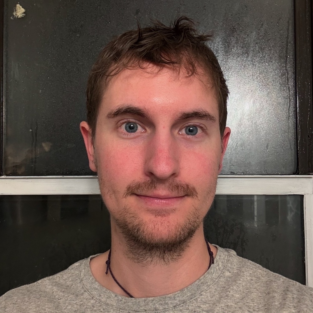 A person with short hair and light facial hair stands facing forward wearing a gray shirt in front of a textured black and frosted window.