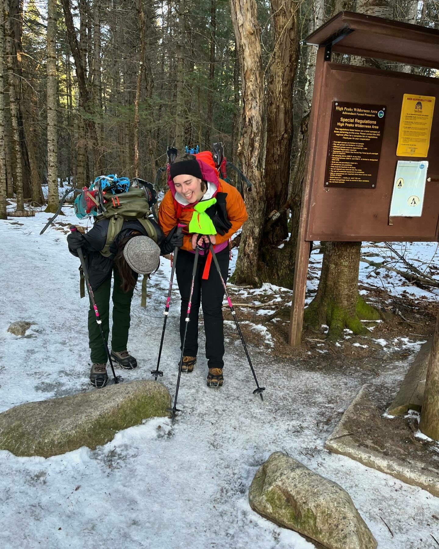 Two hikers with large backpacks stand on a snowy trail, one smiling mid-motion; behind them is a signpost with text, in a wooded area. Signpost text: &ldquo;High Peaks Wilderness Area Special Regulations apply in the High Peaks Wilderness Area&rdquo;