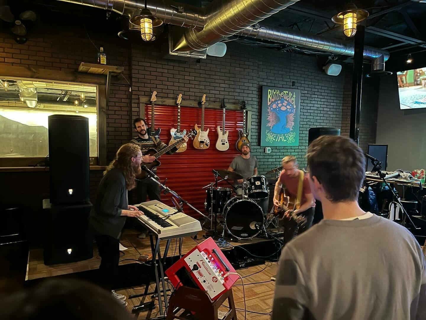A band is performing in a brick-walled bar with guitars on the wall; an audience member watches from the foreground.