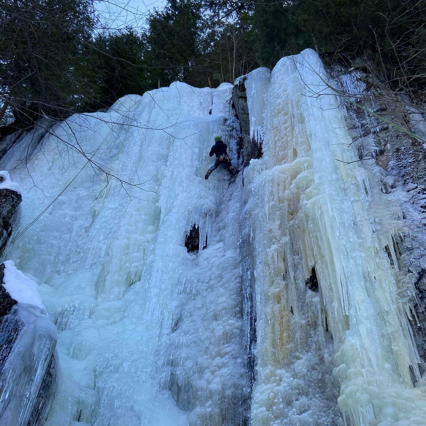 A climber ascends a vertical, frozen waterfall with ice axes, surrounded by snow-covered rocks and trees.