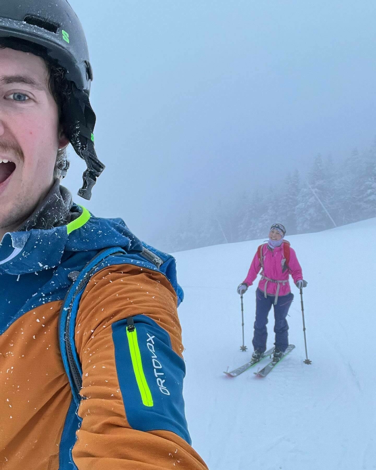 A person in the foreground takes a selfie while another skier stands in the snowy background, both on a misty mountain slope.